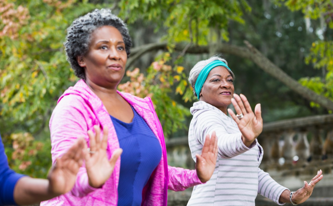 Two senior African American women, doing tai chi exercises at the park.  The focus is on the women on the right, wearing the headband.
