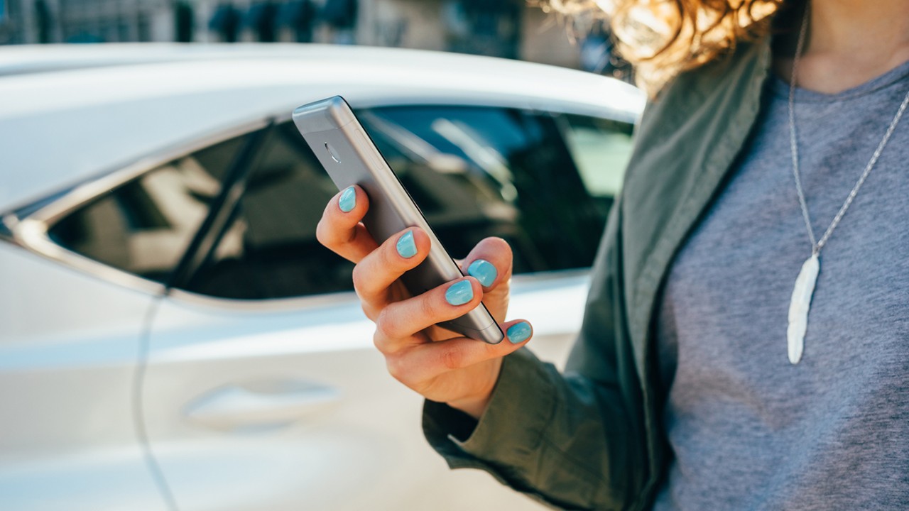 Young woman using smart phone standing on street near road and car. Female's hands holding mobile device in city, close-up.