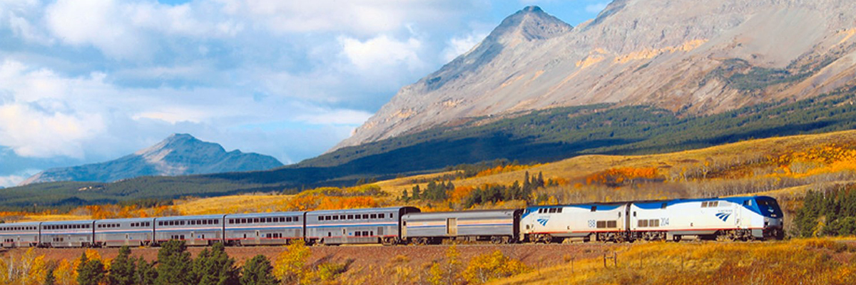 Amtrak train rolling through golden field framed with majestic mountains  