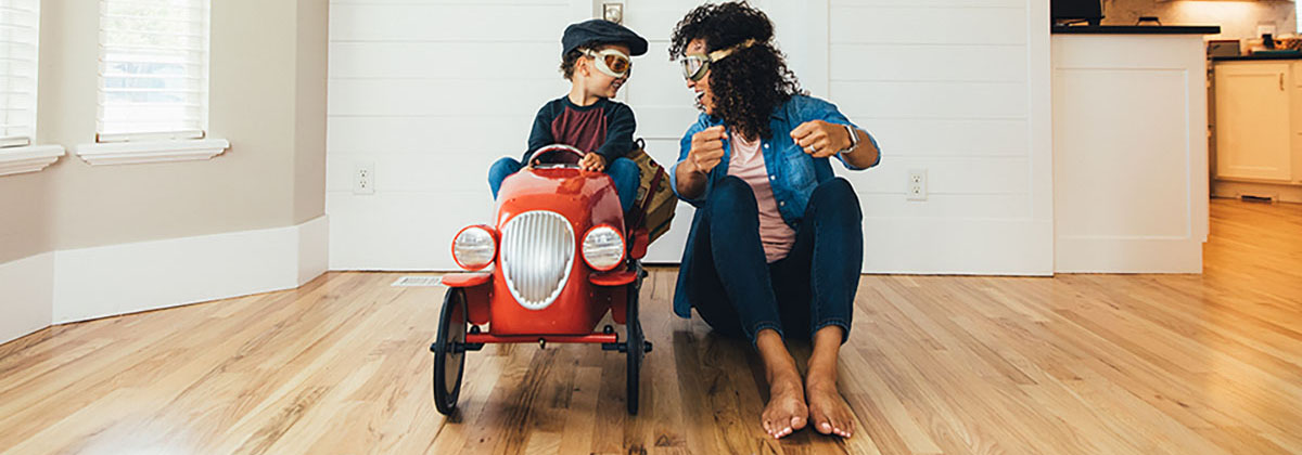 A mother and her young son imagine traveling on the open roads. The boy sits in a red toy car while his mom sits next to him on the floor inside their home. Sometimes a little imagination and a staycation is exactly what a child needs.